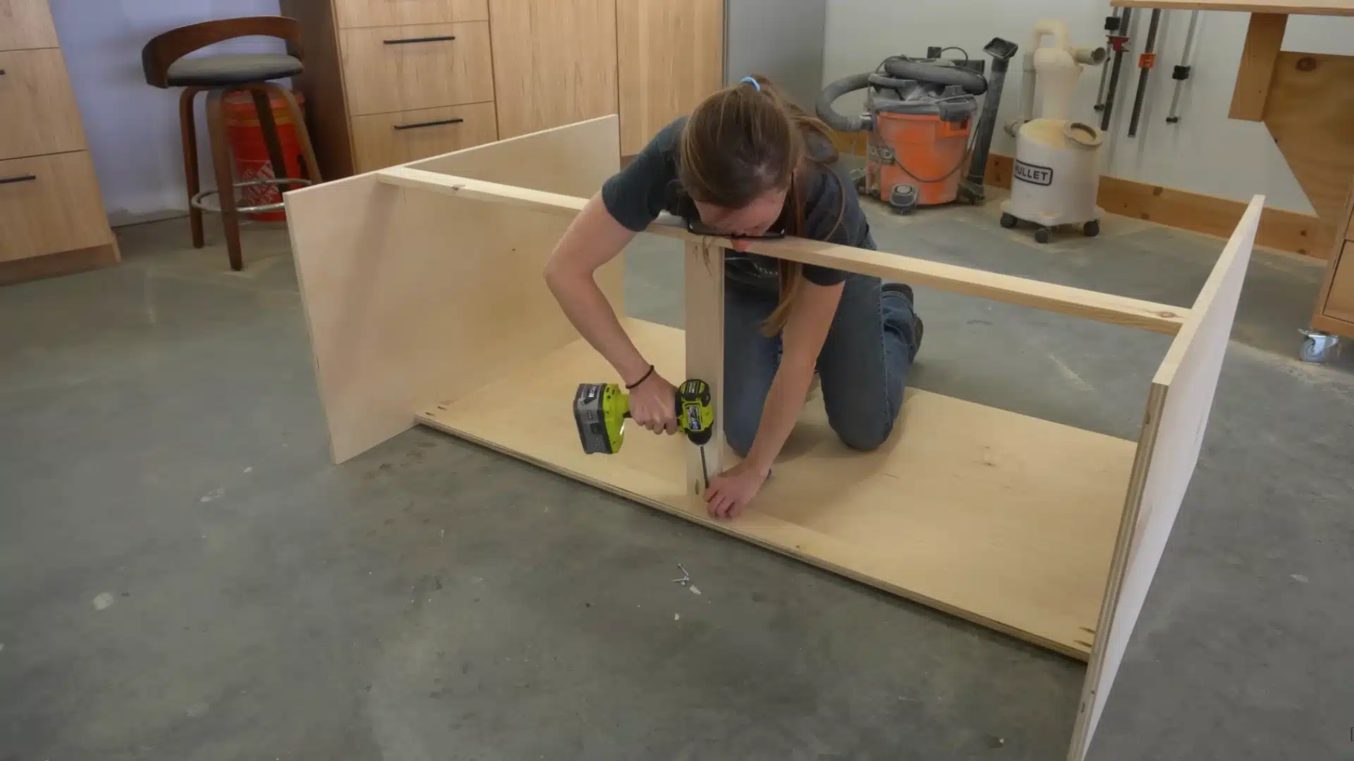 Assembling cabinet frame by securing plywood panels with drill on workshop floor during Murphy bed build